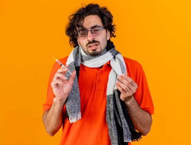 Giovane uomo malato con gli occhiali e sciarpa che tiene la siringa e la fiala con gli occhi chiusi isolati sulla parete arancione