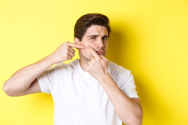 Giovane uomo pop un brufolo sulla guancia, in piedi su sfondo giallo. Concetto di cura della pelle e acne.