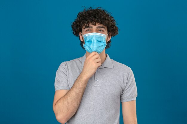 Giovane uomo premuroso in maschera protettiva medica con la mano sul mento sguardo pensieroso in piedi su sfondo blu isolato