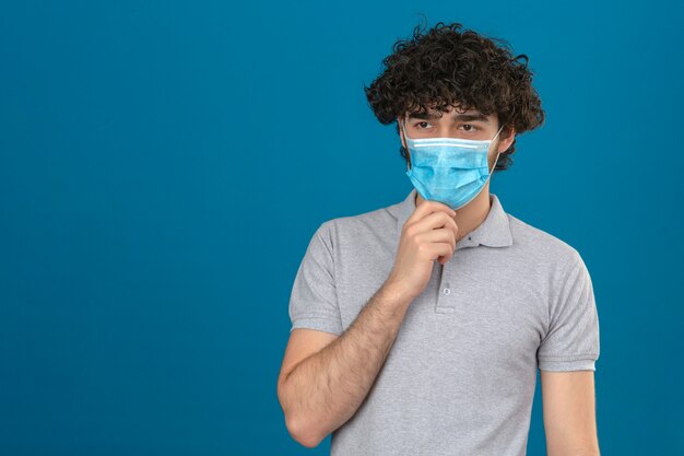 Giovane uomo premuroso in maschera protettiva medica con la mano sul mento sguardo pensieroso in piedi su sfondo blu isolato