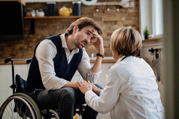 Giovane uomo preoccupato in sedia a rotelle che comunica con un medico mentre lei lo visita a casa