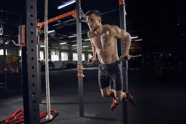 Giovane uomo sano, atleta che fa esercizi, pull-up in palestra. Singolo modello maschile che si esercita duramente e allena la parte superiore del corpo. Concetto di stile di vita sano, sport, fitness, bodybuilding, benessere.