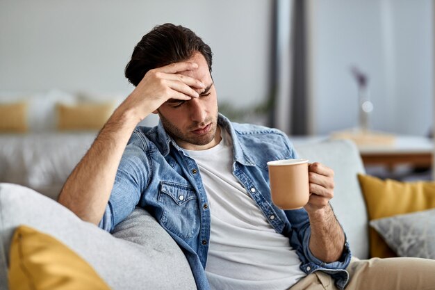 Giovane uomo sconvolto che beve caffè e pensa a qualcosa mentre è seduto sul divano