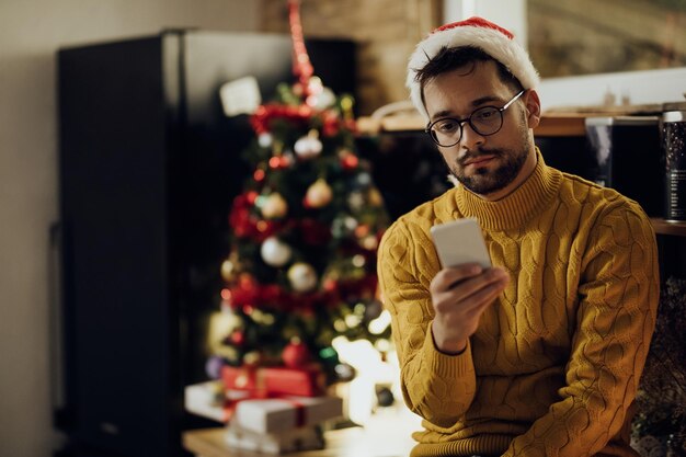 Giovane uomo sconvolto che legge un messaggio di testo sul cellulare mentre è solo a casa a Natale