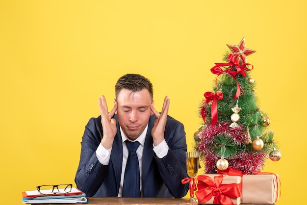 giovane uomo seduto al tavolo vicino albero di Natale e regali su giallo