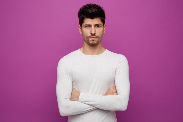 Giovane uomo serio e bello che guarda l'obbiettivo tenendo le braccia incrociate su sfondo viola