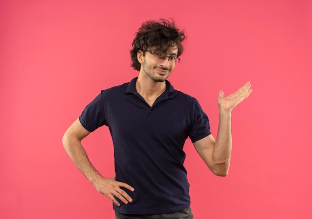 Giovane uomo soddisfatto in camicia nera con occhiali ottici finge di tenere qualcosa a portata di mano e mette la mano sulla vita isolata sul muro rosa