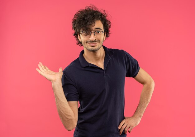 Giovane uomo soddisfatto in camicia nera con vetri ottici alza la mano fingendo di tenere qualcosa di isolato sul muro rosa