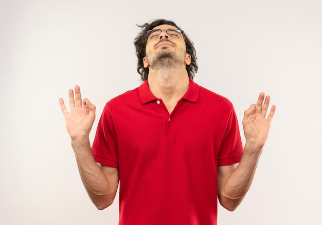 Giovane uomo soddisfatto in camicia rossa con occhiali ottici gesti il segno giusto della mano e finge di meditare isolato sul muro bianco