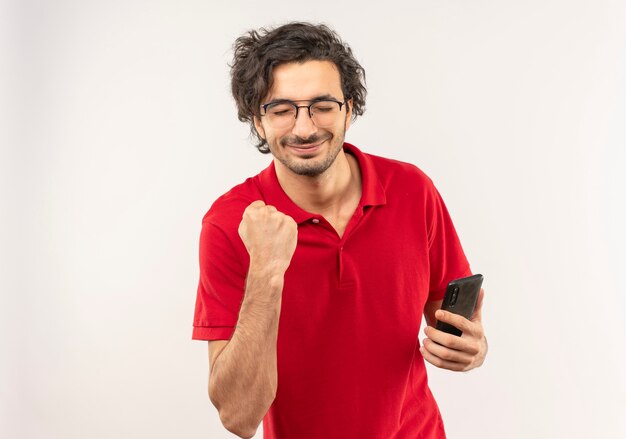 Giovane uomo soddisfatto in camicia rossa con vetri ottici tiene il telefono e tiene il pugno isolato sul muro bianco con copia spac