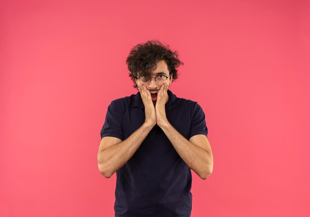 Giovane uomo sorpreso in camicia nera con occhiali ottici mette le mani sul viso isolato sul muro rosa