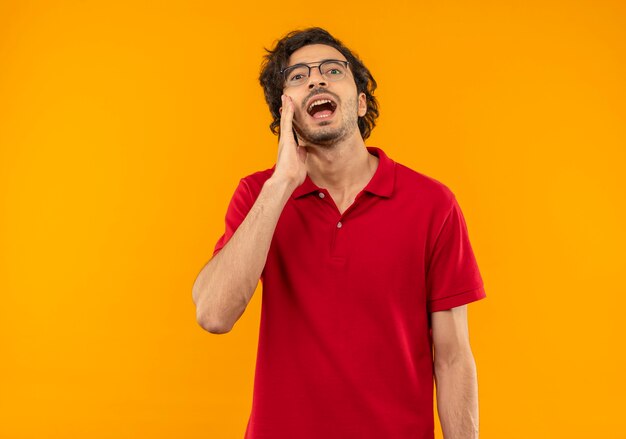 Giovane uomo sorpreso in camicia rossa con occhiali ottici mette la mano sul viso e finge di chiamare qualcuno isolato sulla parete arancione