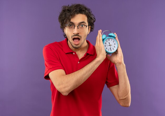 Giovane uomo sorpreso in camicia rossa con vetri ottici tiene l'orologio con entrambe le mani e sembra isolato sulla parete viola