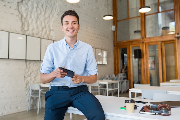 Giovane uomo sorridente alla moda in ufficio di co-working, libero professionista di avvio che tiene facendo uso della compressa