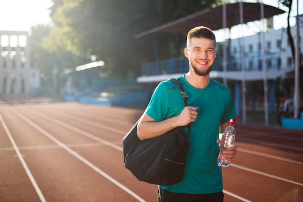 Giovane uomo sorridente che guarda felicemente a porte chiuse con borsa sportiva sulla spalla e bottiglia di acqua pura in mano sulla pista da corsa dello stadio