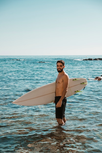 Giovane uomo sorridente con tavola da surf vicino alla spiaggia in acqua