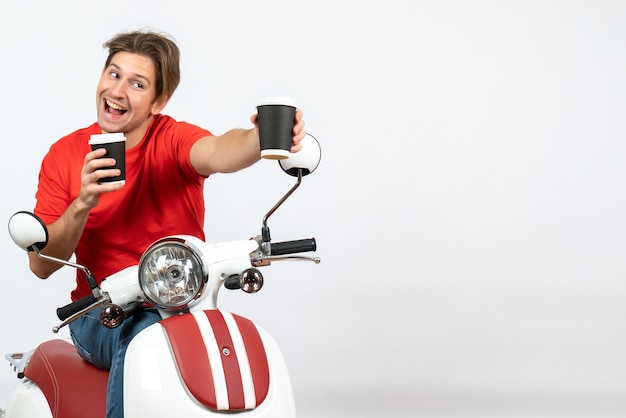 Giovane uomo sorridente del corriere in uniforme rossa che si siede sul motorino che dà uno dei bicchieri di carta sulla parete gialla