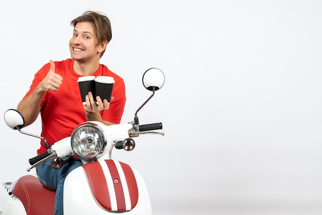 Giovane uomo sorridente del corriere in uniforme rossa che si siede sul motorino che tiene i bicchieri di carta che fa il gesto giusto sulla parete gialla