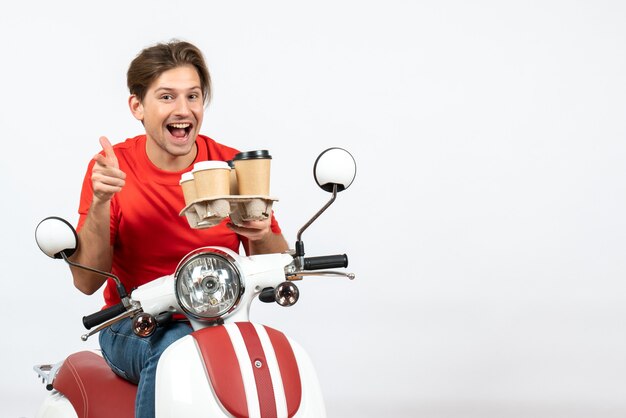 Giovane uomo sorridente del corriere in uniforme rossa che si siede sul motorino e che tiene gli ordini che indicano in avanti sulla parete gialla