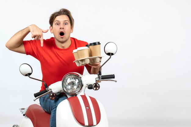 Giovane uomo sorridente del corriere in uniforme rossa che si siede sullo scooter e che tiene gli ordini rivolti verso il basso sulla parete gialla