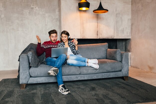 Giovane uomo sorridente e donna seduta a casa in inverno guardando nel laptop con espressione del viso scioccato sorpreso, utilizzando internet, coppia nel tempo libero insieme, emozione felice e positiva