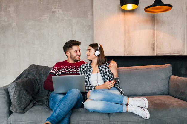 Giovane uomo sorridente e donna seduta a casa in inverno, lavorando al computer portatile, tenendo lo smartphone, ascoltando le cuffie, coppia nel tempo libero che trascorre del tempo online, libero professionista, incontri