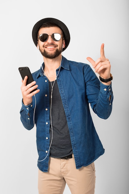 Giovane uomo sorridente felice bello che ascolta la musica in auricolari isolati su sfondo bianco studio, tenendo lo smartphone, indossa una camicia di jeans, cappello e occhiali da sole