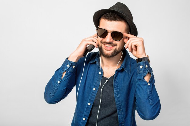 Giovane uomo sorridente felice bello che ascolta la musica in auricolari isolati su sfondo bianco studio, tenendo lo smartphone, indossa una camicia di jeans, cappello e occhiali da sole