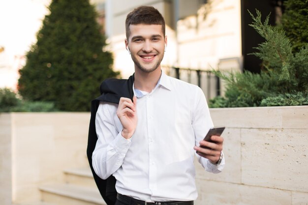 Giovane uomo sorridente in camicia bianca con auricolari wireless che tiene una giacca nera sulla spalla con il cellulare in mano mentre guarda con gioia nella fotocamera all'aperto