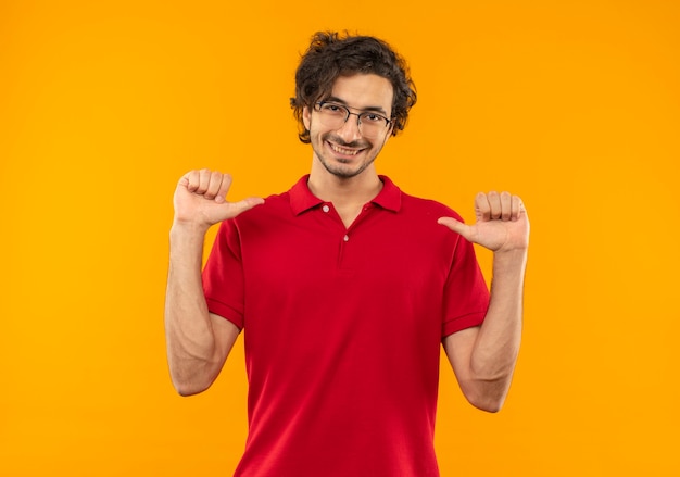 Giovane uomo sorridente in camicia rossa con occhiali ottici punta a se stesso isolato sulla parete arancione