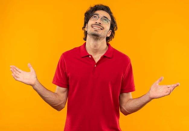 Giovane uomo sorridente in camicia rossa con vetri ottici tiene le mani aperte e guarda in alto isolato sulla parete arancione