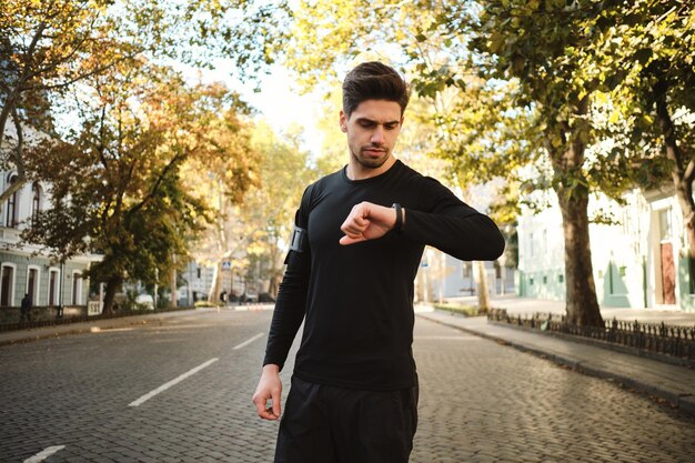 Giovane uomo sportivo attraente che controlla premurosamente il suo orologio dopo la corsa mattutina sulla strada della città