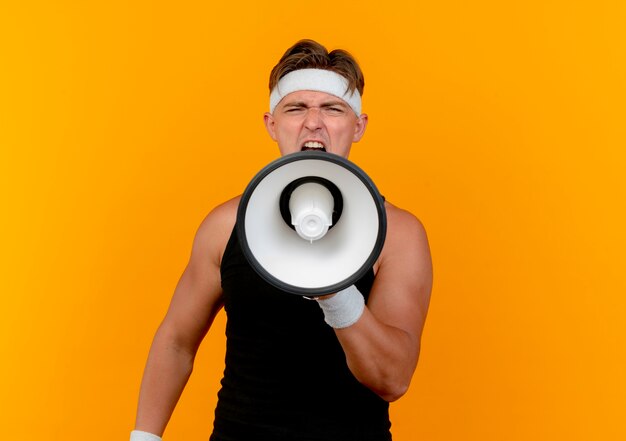 Giovane uomo sportivo bello che indossa la fascia e braccialetti che grida in altoparlante alla macchina fotografica isolata su priorità bassa arancione con lo spazio della copia