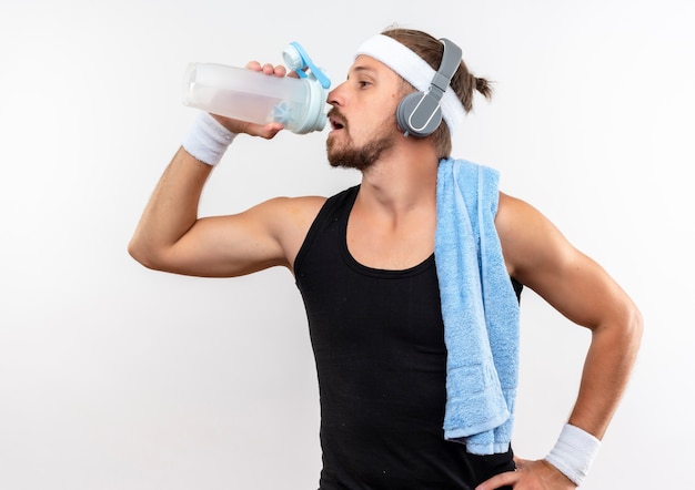 Giovane uomo sportivo bello che indossa la fascia e braccialetti e cuffie che bevono acqua dalla bottiglia di acqua con l'asciugamano sulla spalla e la mano sulla vita isolata su spazio bianco