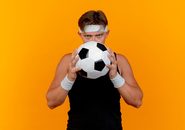 Giovane uomo sportivo bello che indossa la fascia e polsini che tengono e che guarda da dietro il pallone da calcio alla macchina fotografica isolata su fondo arancio