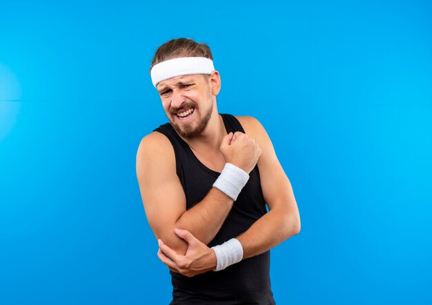 Giovane uomo sportivo bello dolorante che indossa fascia e braccialetti stringendo il pugno e tenendo il gomito isolato sulla parete blu con spazio di copia