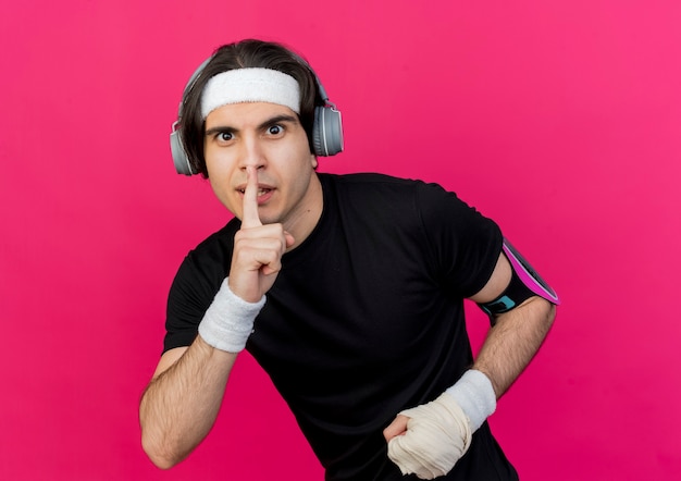 Giovane uomo sportivo che indossa abbigliamento sportivo e fascia con auricolari e fascia da braccio dello smartphone che fa gesto di silenzio con il dito sulle labbra