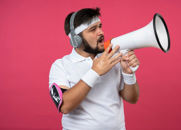 Giovane uomo sportivo che indossa la fascia e il braccialetto con le cuffie e la fascia da braccio del telefono parla sull'altoparlante