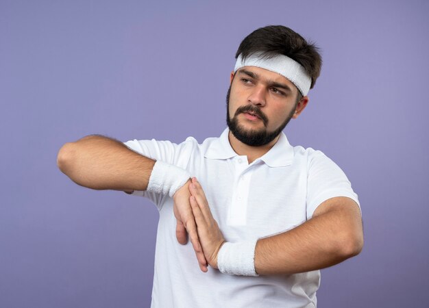 Giovane uomo sportivo guardando al lato che indossa la fascia e il braccialetto che tengono le mani insieme isolato sulla parete verde