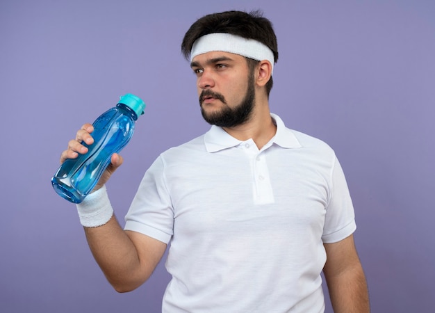 Giovane uomo sportivo guardando lato indossando la fascia e il braccialetto tenendo la bottiglia d'acqua isolata sul verde