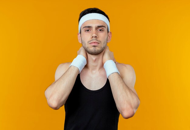 Giovane uomo sportivo in fascia con la faccia seria che tocca il suo collo in piedi sopra la parete arancione