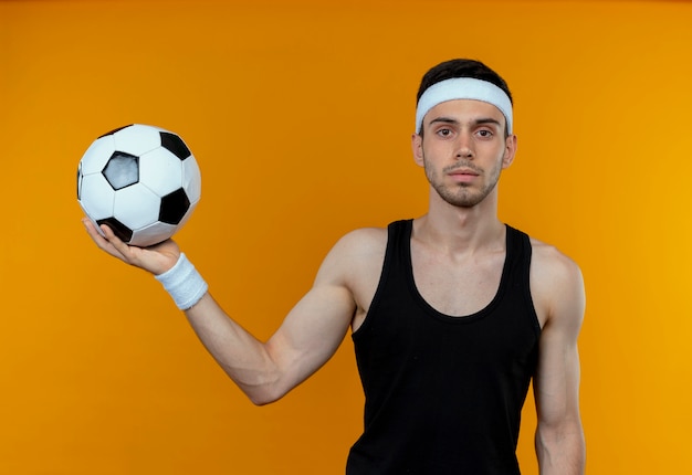 Giovane uomo sportivo in fascia tenendo il pallone da calcio con sicura espressione seria in piedi sopra la parete arancione
