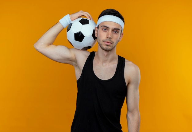 Giovane uomo sportivo in fascia tenendo il pallone da calcio sulla sua spalla con espressione seria fiduciosa in piedi sopra la parete arancione