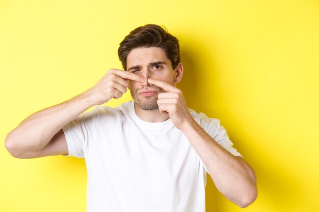 Giovane uomo spremitura brufolo sul naso, in piedi su sfondo giallo. Concetto di cura della pelle e acne.