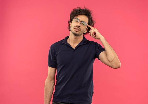 Giovane uomo stanco in camicia nera con occhiali ottici mette il dito sulla testa isolata sul muro rosa