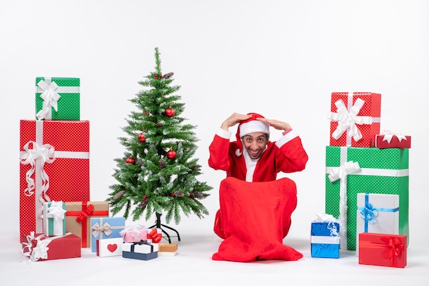 Giovane uomo vestito da Babbo Natale con doni e albero di Natale decorato seduto per terra mettendo entrambe le mani