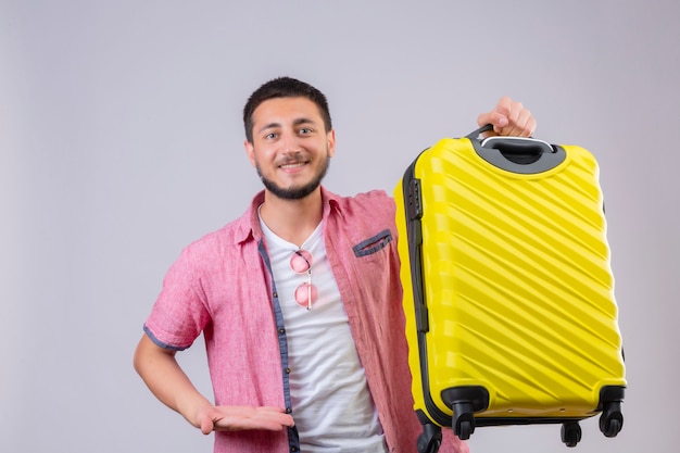 Giovane valigia bella della tenuta del tipo del viaggiatore che esamina macchina fotografica con il fondo bianco controllante positivo e felice di sorriso sicuro