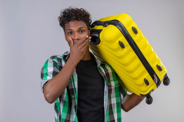 Giovane viaggiatore afroamericano uomo che tiene la valigia sorpreso e stupito guardando la telecamera che copre la bocca con la mano in piedi su sfondo bianco
