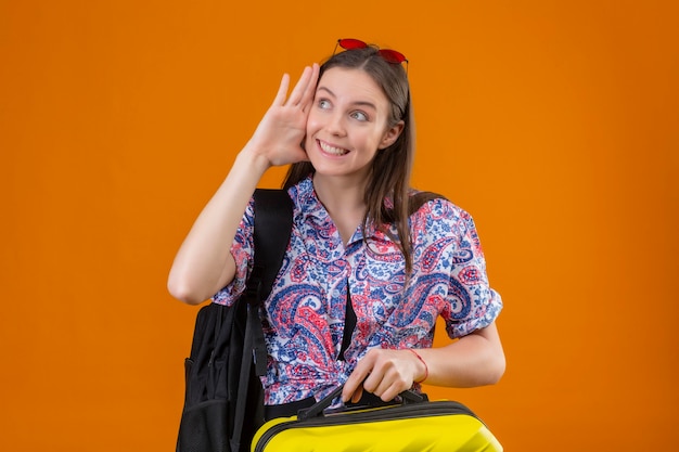 Giovane viaggiatore donna che indossa gli occhiali da sole rossi sulla testa in piedi con uno zaino tenendo la valigia guardando a parte sorridendo allegramente tenendo la mano vicino al suo orecchio cercando di ascoltare la conversazione di som