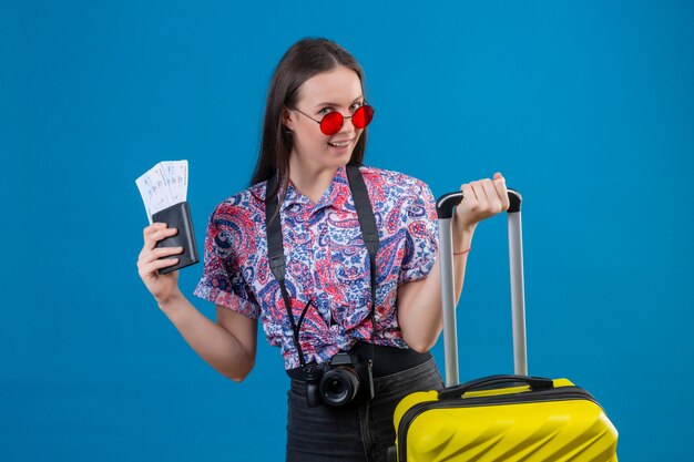 Giovane viaggiatore donna che indossa occhiali da sole rossi in piedi con la valigia gialla tenendo il passaporto e biglietti sorridendo allegramente guardando la fotocamera con la faccia felice su sfondo blu